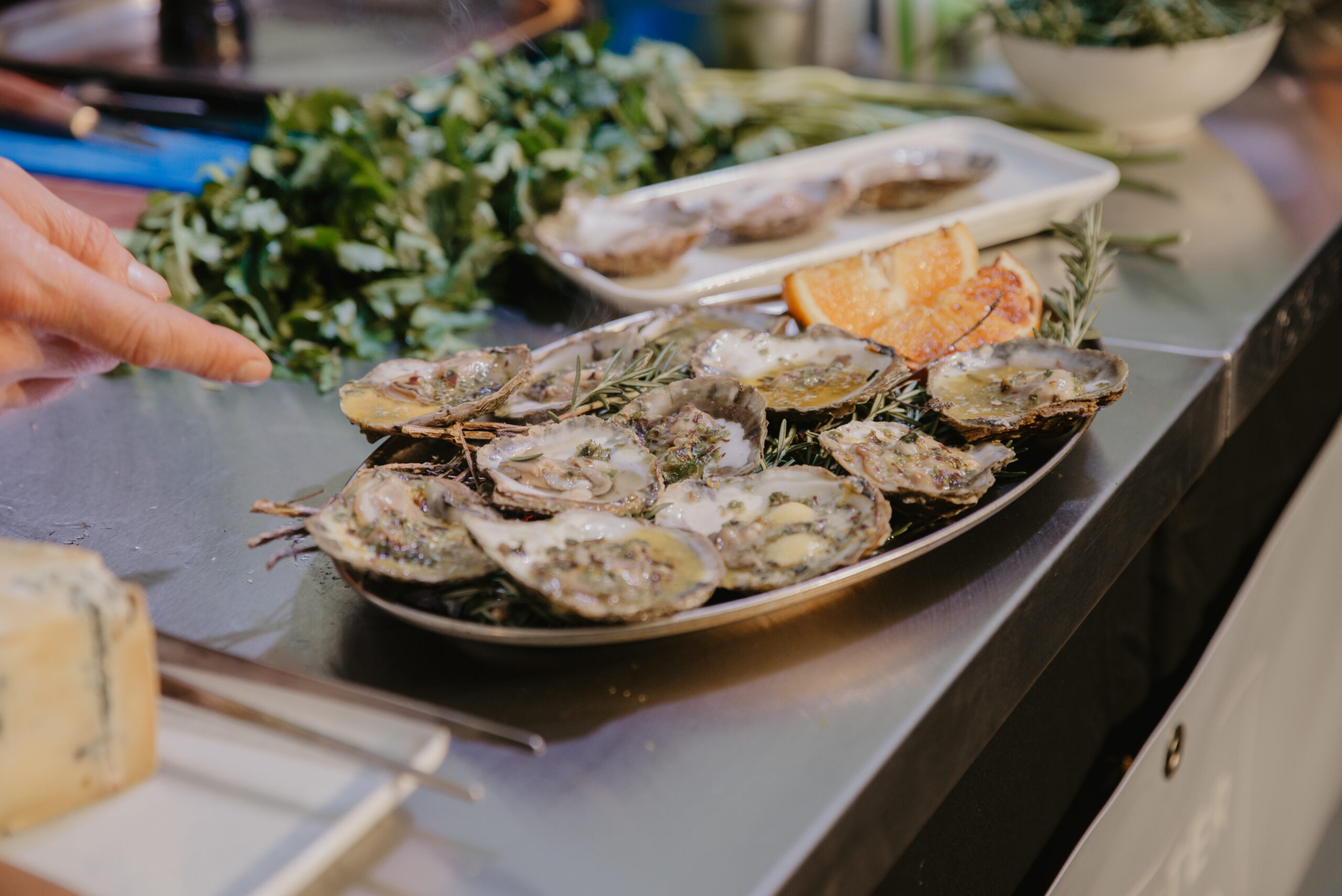A large platter of fresh oysters sits on a counter, garnished with lemon and herbs, while a hand reaches toward the seafood.