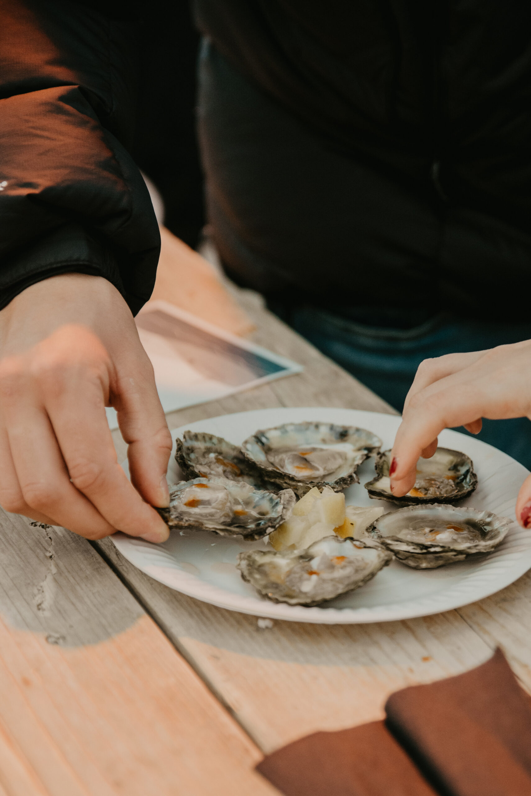 Hands served oysters on a white plate at a table, with lemon wedges and a teal napkin