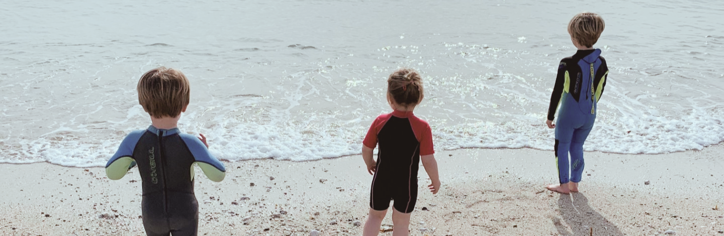 Three children in wetsuits at the water's edge on Gyllyngvase Beach in Falmouth, Cornwall