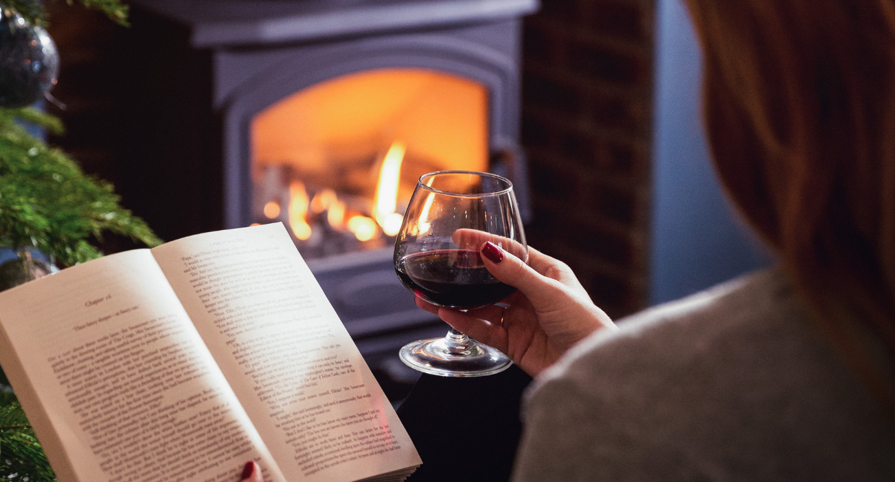 A person relaxing by a fireplace, holding a glass of red wine while reading a book in a cosy, softly lit setting.